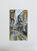 A watercolor painting of a narrow historic street lined with timber-framed buildings, shown centered within a wide white mat or mount.