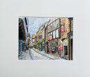 A matted art print featuring a hand-painted watercolor street scene of a historic town with brick buildings, shopfronts, and a cobblestone street under a blue sky.