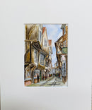 A matted watercolor painting of York’s Shambles - a historic street lined with timber-framed buildings under a pale blue sky.