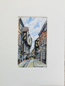 A matted watercolor painting of a narrow historic street with timber-framed buildings, a blue sky, and a few birds, shown against a white mat board.
