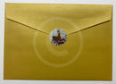 A  gold envelope with a textured metallic finish, featuring a circular custom illustration sticker of a bicycle and figure on the front flap, set against a plain white background.
