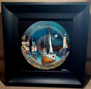 Framed artwork of a coastal scene with a boat, houses, and a lighthouse.