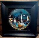 Framed artwork of a coastal scene with a boat, houses, and a lighthouse.