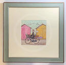 Meet the Mustards: Out For a Bit of Brunch by Sam Toft, mounted miniature print. A man rides on a bicycle with his dog sat in the front basket, they have a picnic basket on the back. They are riding past two pink and yellow beach huts. Framed in Nordic Blue. 