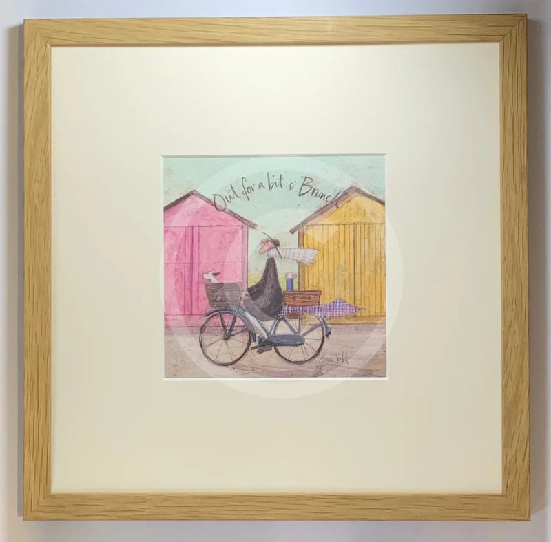 Meet the Mustards: Out For a Bit of Brunch by Sam Toft, mounted miniature print. A man rides on a bicycle with his dog sat in the front basket, they have a picnic basket on the back. They are riding past two pink and yellow beach huts. Framed in Oak. 