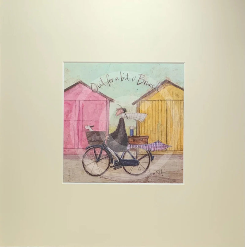 Meet the Mustards: Out For a Bit of Brunch by Sam Toft, mounted miniature print. A man rides on a bicycle with his dog sat in the front basket, they have a picnic basket on the back. They are riding past two pink and yellow beach huts. 