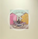 Meet the Mustards: Out For a Bit of Brunch by Sam Toft, mounted miniature print. A man rides on a bicycle with his dog sat in the front basket, they have a picnic basket on the back. They are riding past two pink and yellow beach huts. 