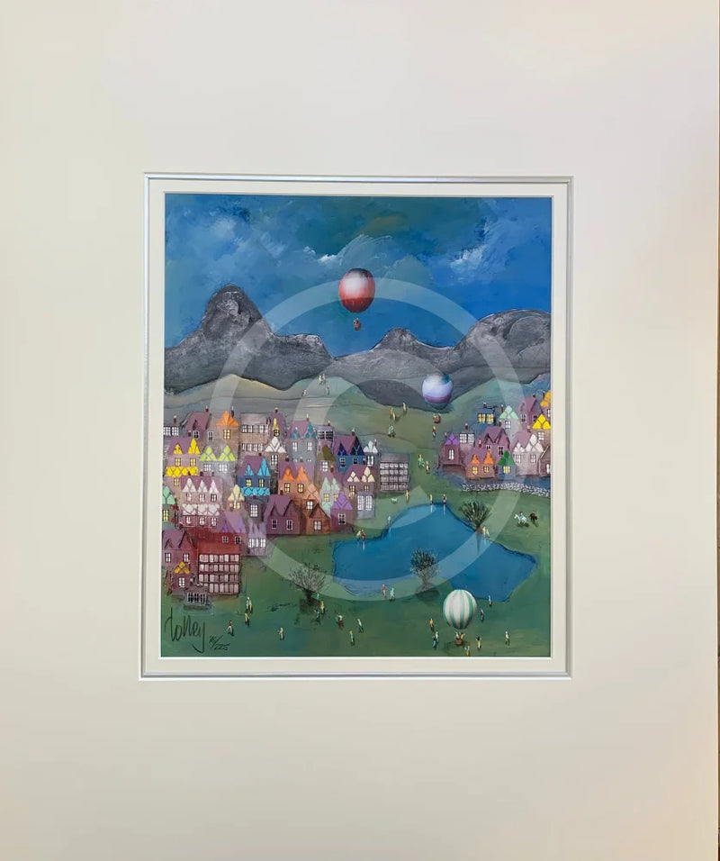 Mounted artwork of a colourful village scene with balloons against a mountainous background.