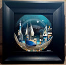 Framed artwork of a lighthouse, sailboat, and houses on a moon-shaped platform.